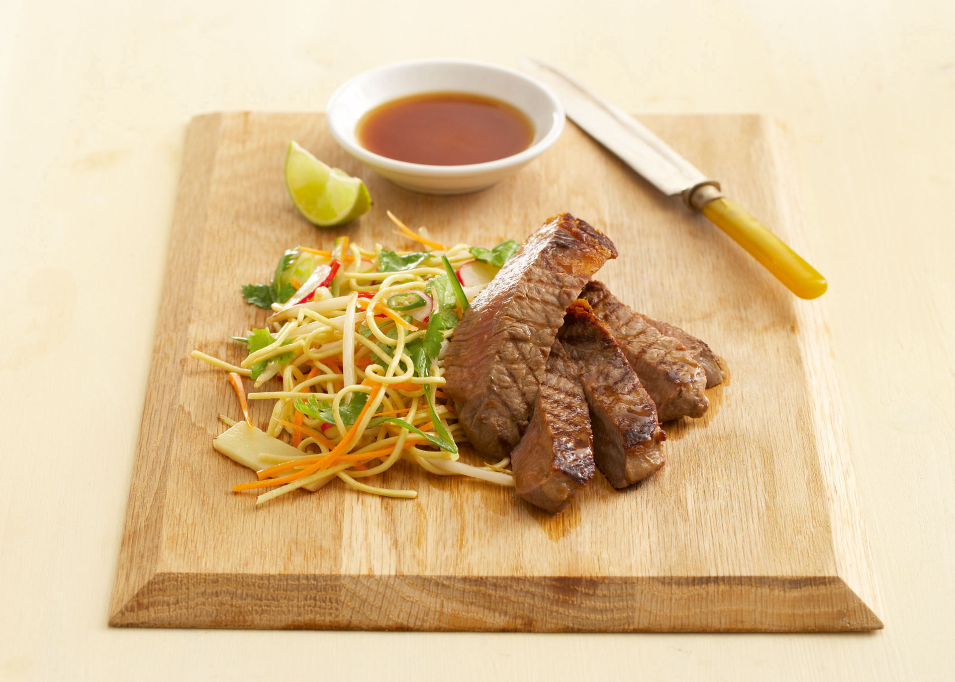 Eiernudel-Salat, gegrilltes Steak in Scheiben und ein Schälchen mit Sauce angerichtet auf einem Holzbrett
