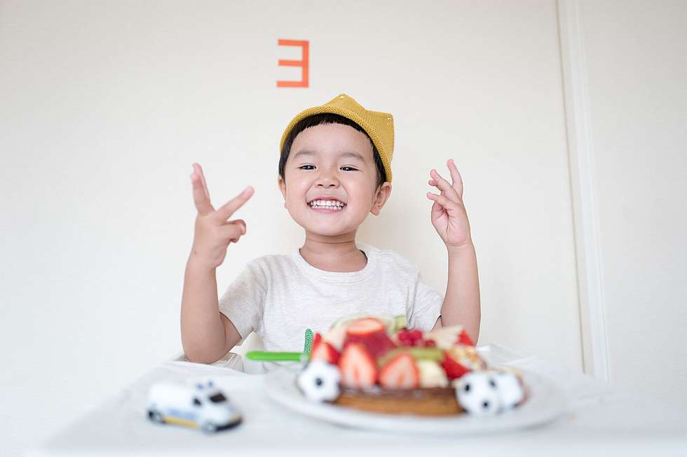 3. Geburtstag: Kleiner Junge sitzt am Tisch und hält 3 Finger hoch