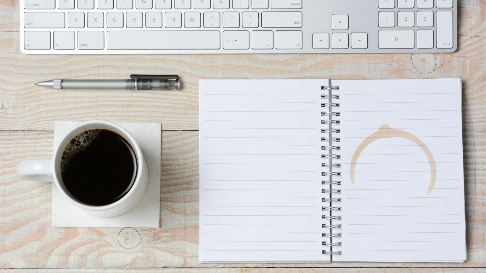 Draufsicht auf einen Schreibtisch, auf dem eine Kaffeetasse, ein Notizblock und eine Tastatur liegen.