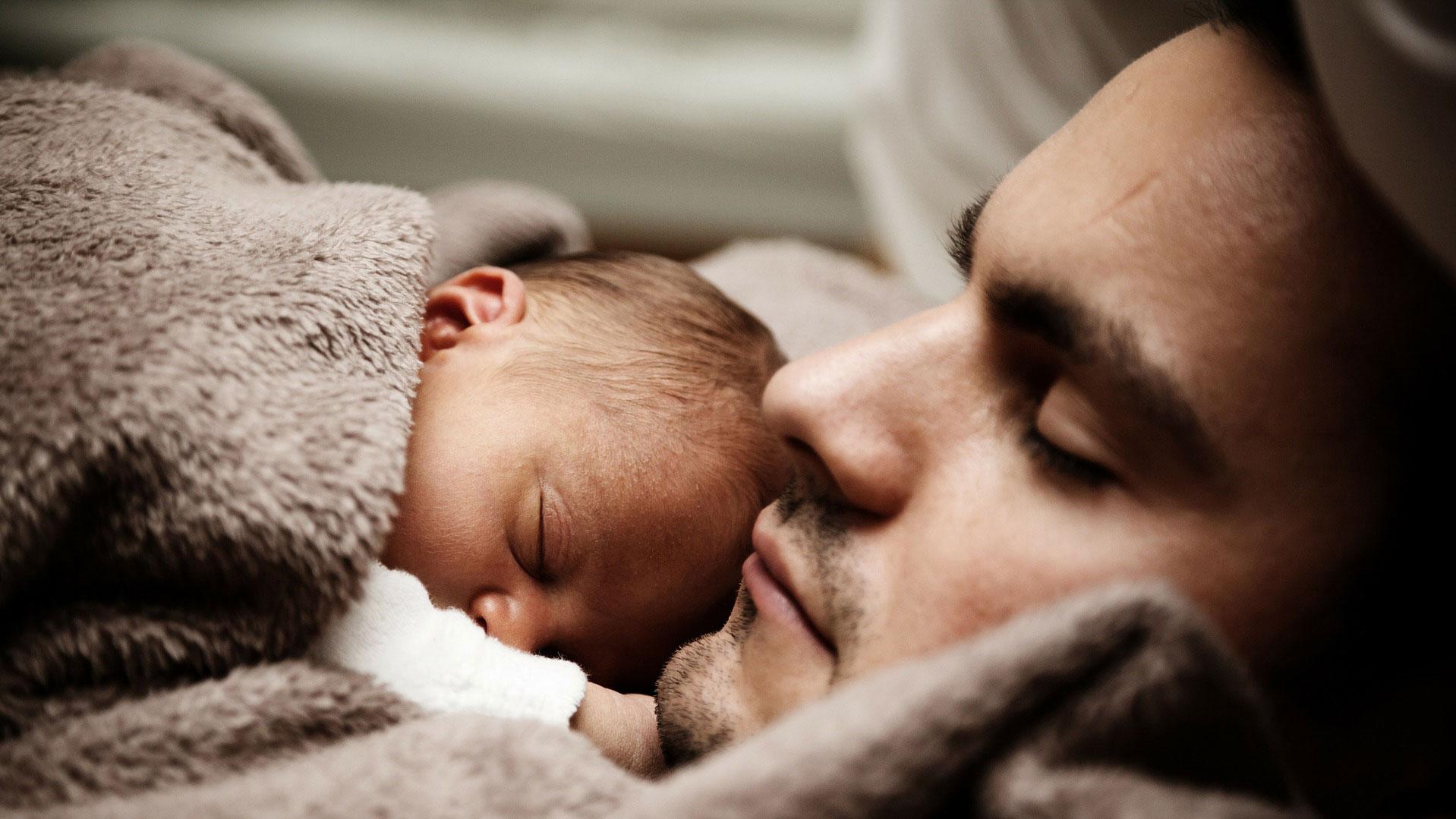 Junger Vater liegt mit Säugling auf der Brust unter einer Decke 
