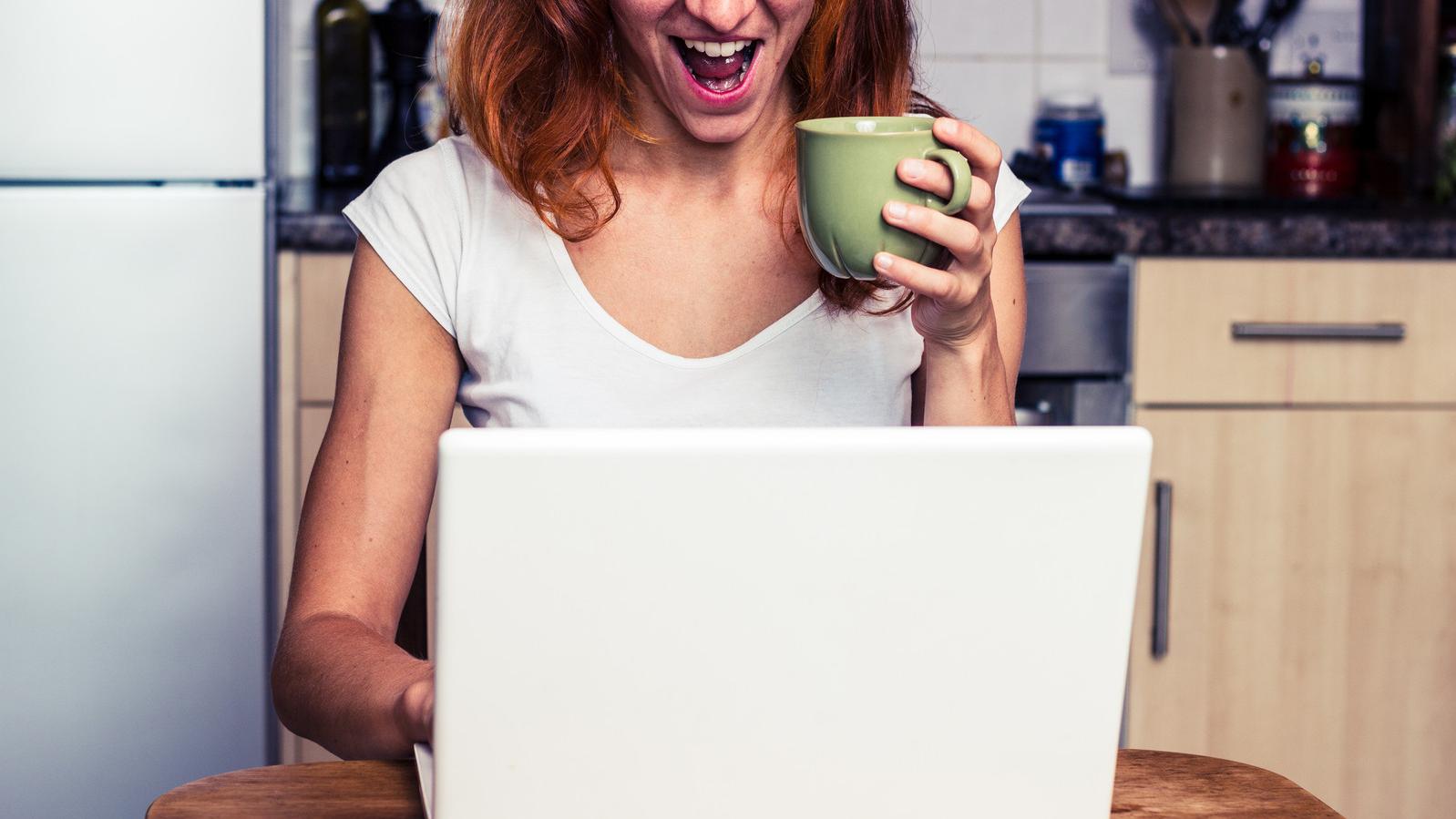 Frau sitzt mit einem Kaffee in der Hand vor einem Laptop am Küchentisch und lacht mit offenem Mund