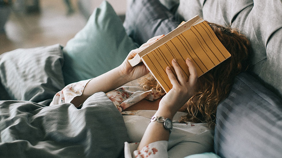 Ruhe genießen: Frau liegt unter einer Decke auf der Couch und versteckt ihr Gesicht hinter einem Buch.