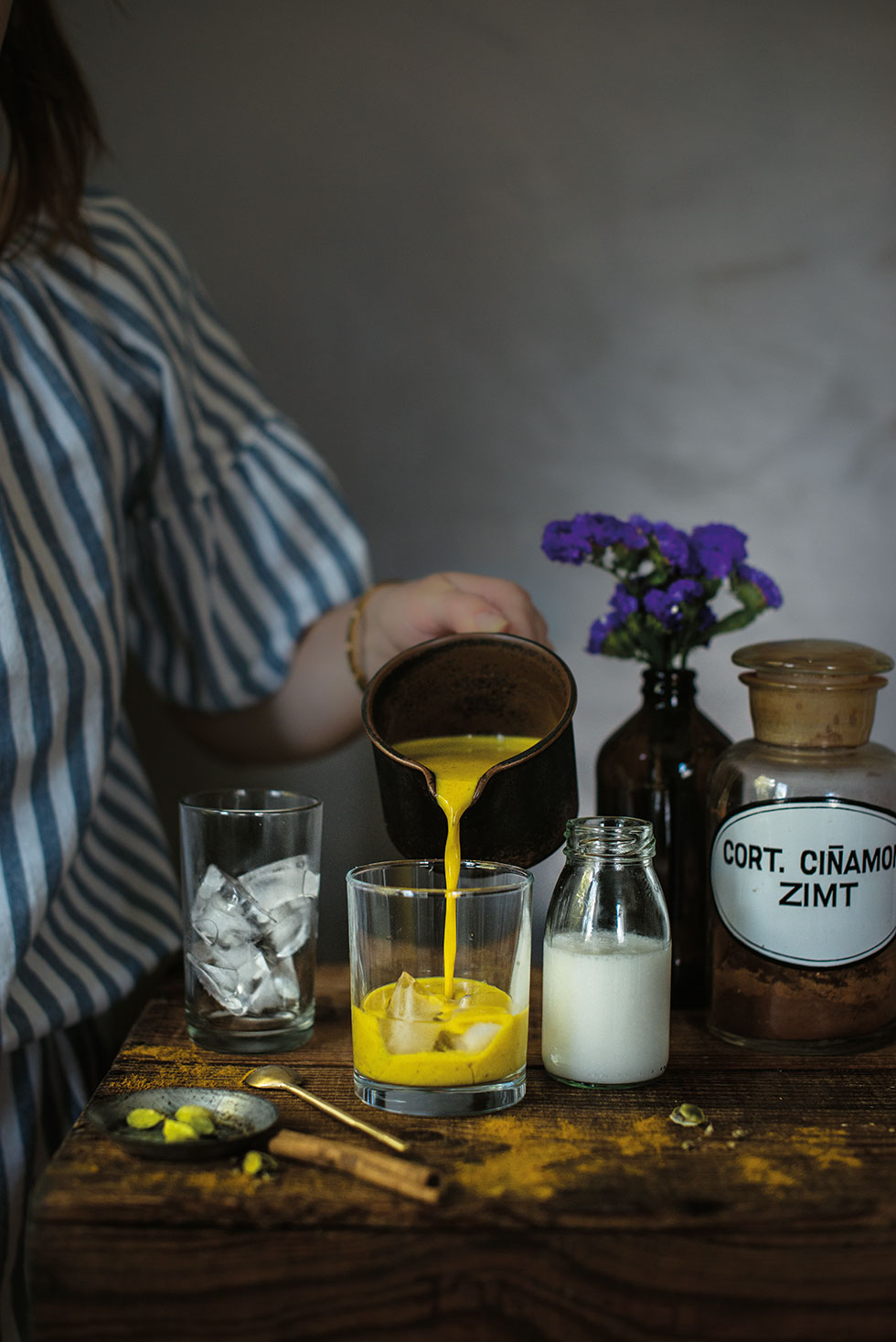 Rezeptbild: Geeiste Golden Milk wird aus Kännchen in ein Glas mit Eiswürfel gefüllt, daneben weiter Zutaten wie Mandelmilch, Kardamomkapseln und Zimt