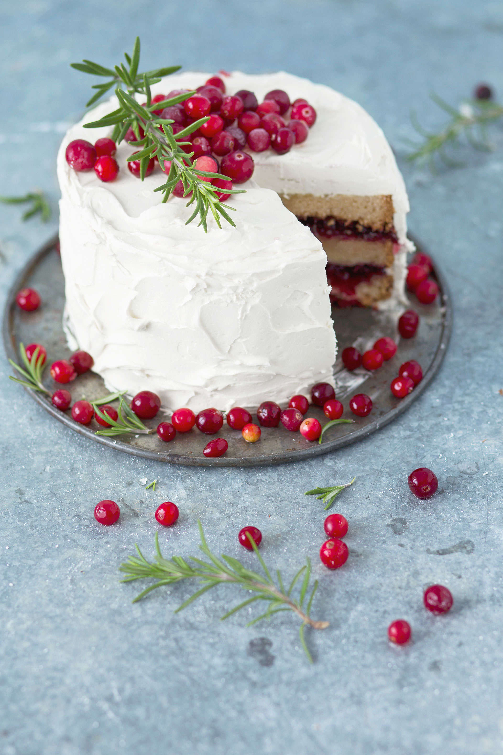Angeschnittene Torte mit Cranberry-Füllung und Frosting, dekoriert mit frischen Cranberries und Rosmarin