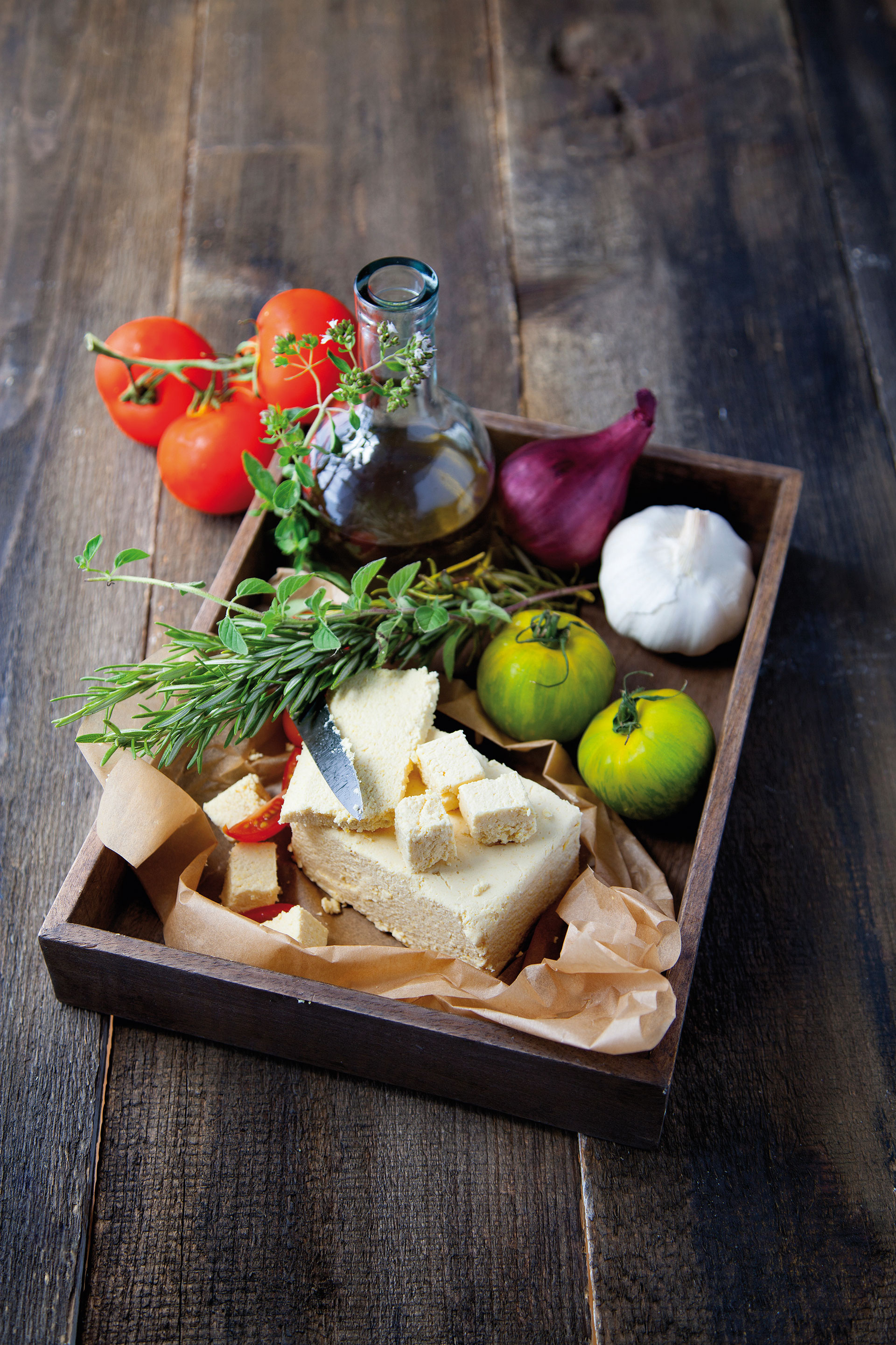 Tablett mit einem Block veganem Käse nach Feta-Art und weiteren Zutaten (Kräuterzweige, Öl, Tomaten, Zwiebel, Knoblauch)