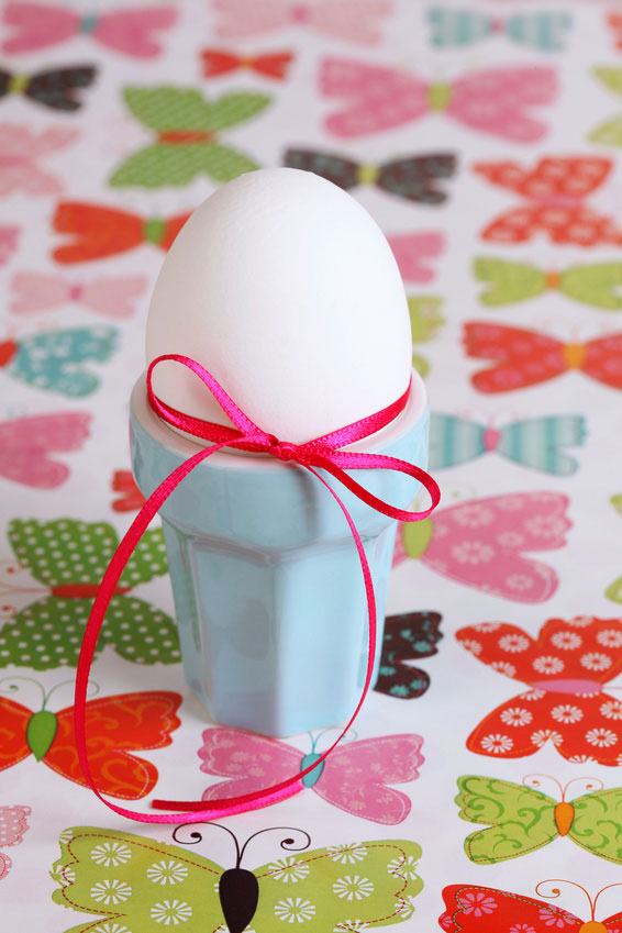 Bild mit einem weißen Ei in einem hellblauen Eierbecher und pinker Schleifer auf buntem Tisch.