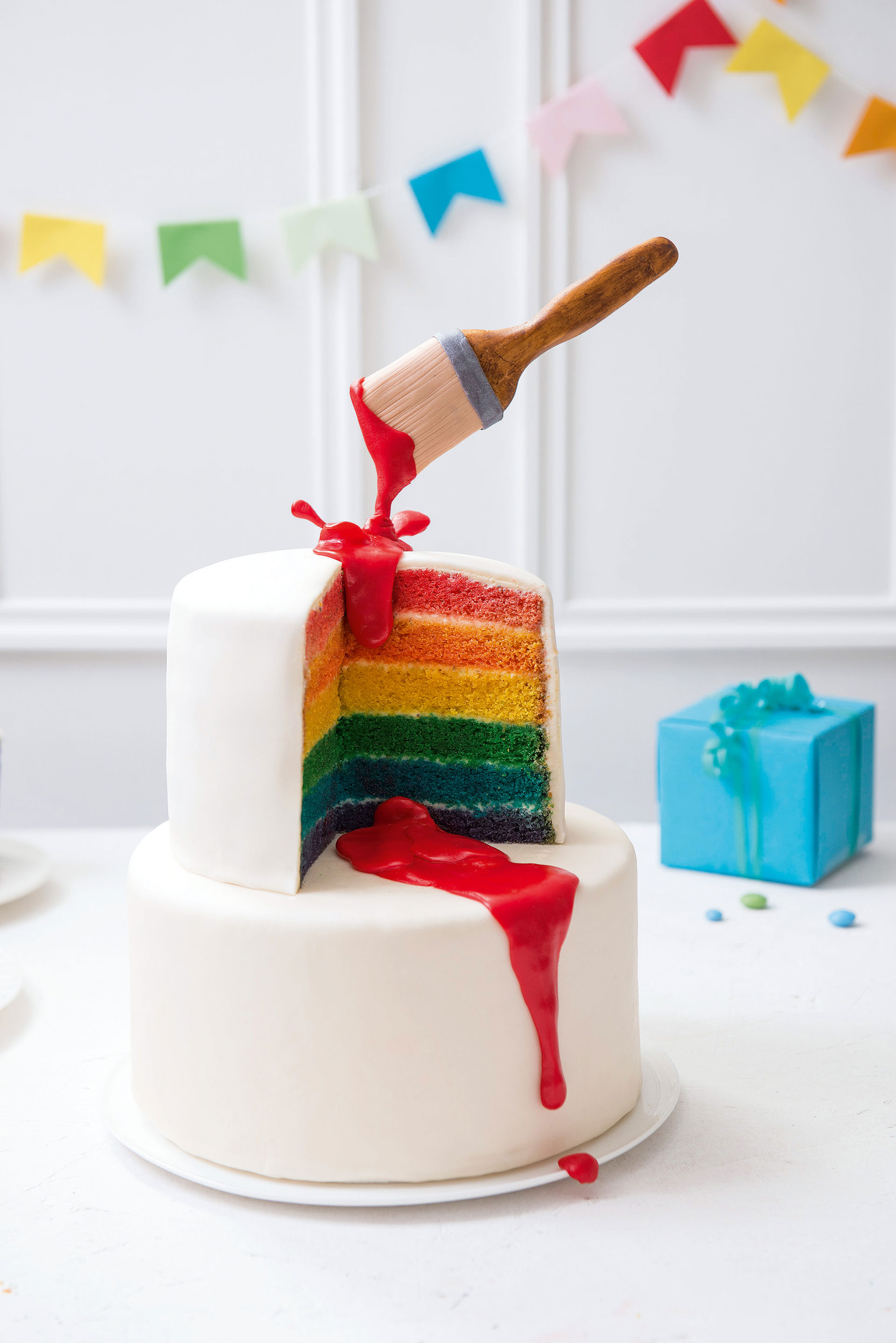 Kuchen mit Schichten in Regenbogenfarben und weißem Frosting, dekoriert mit einem oben am Kuchen befestigten Pinsel aus Fondant